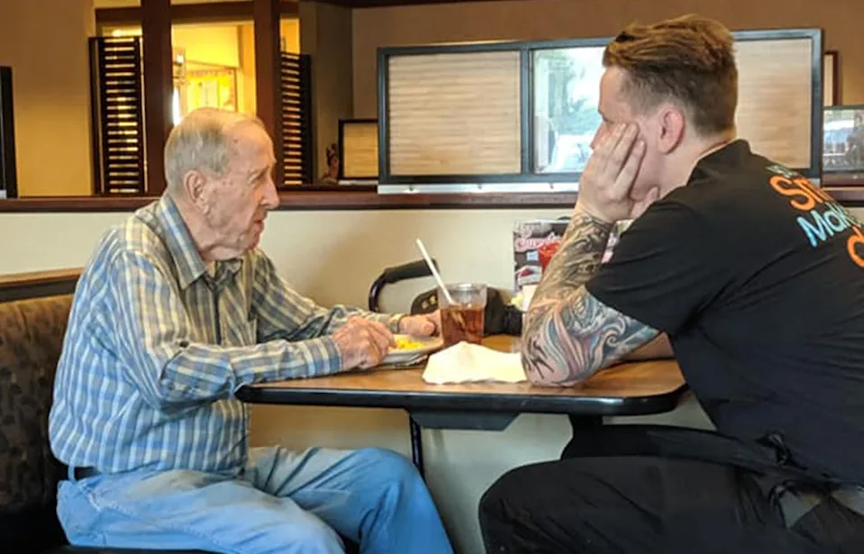 Waiter sitting beside elderly veteran at diner booth, talking warmly.
