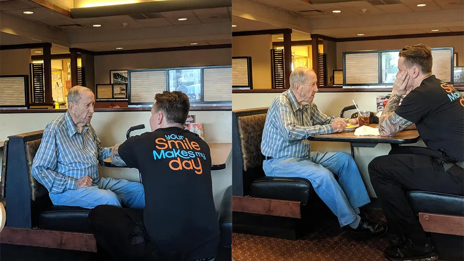 Waiter kneels beside elderly veteran at a diner table, smiling as they talk.